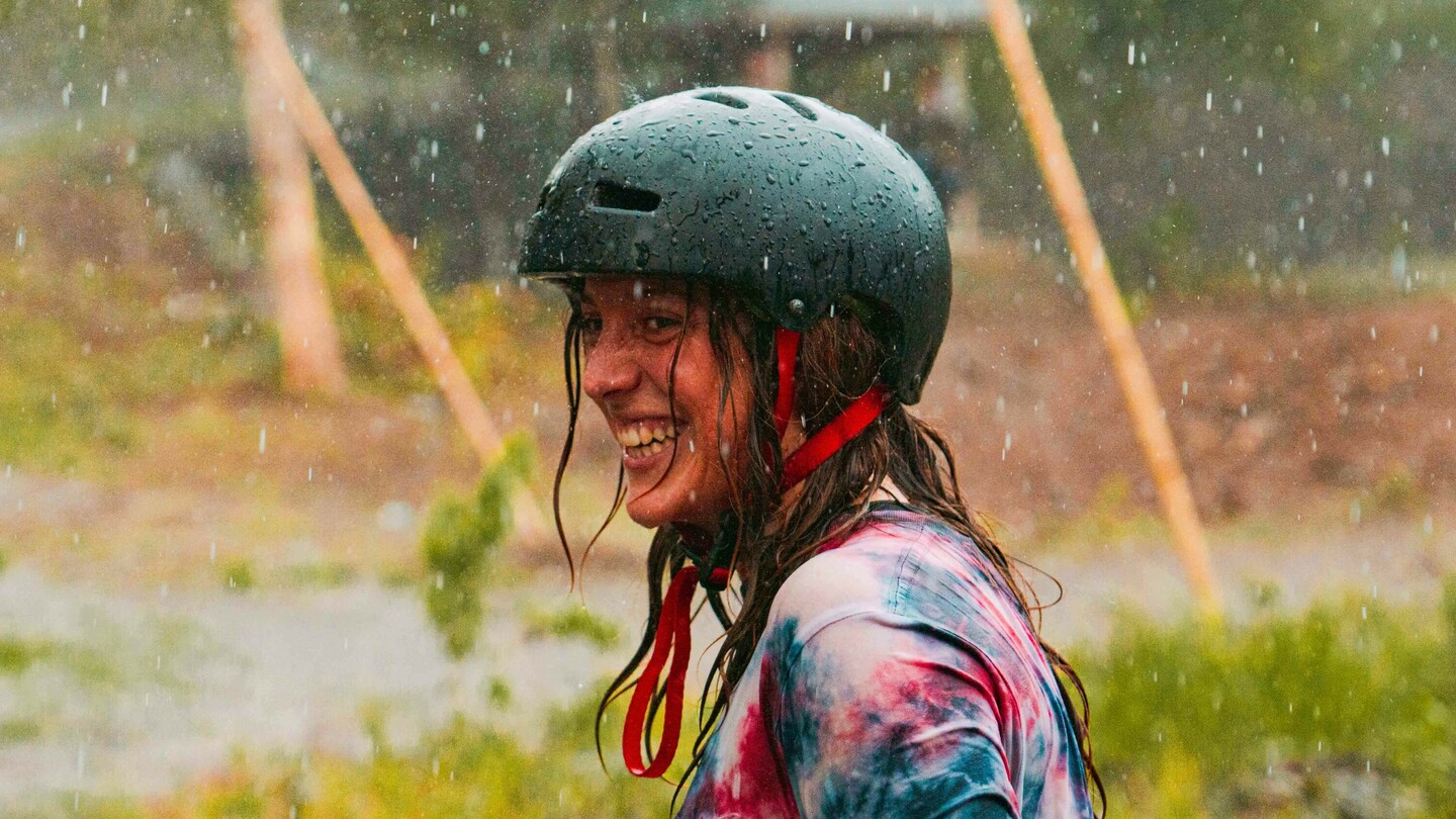 Anna Pixner, a downhill skateboarder, is smiling in the rain wearing a black helmet and a colorful tie-dye T-shirt. The background shows a rural setting with a pavilion and several people.