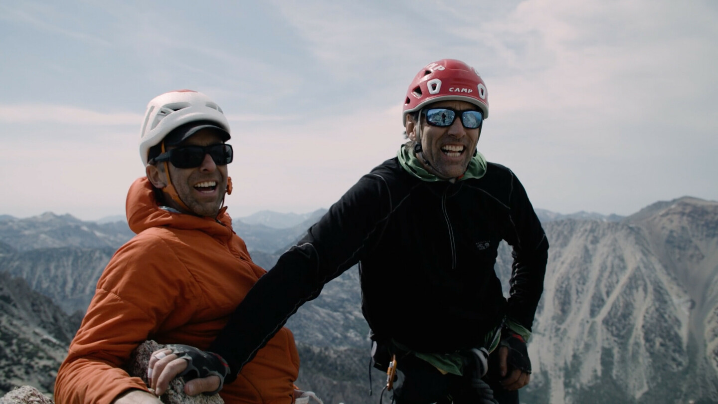 On the left stands Timmy O’Neill wearing an orange jacket and helmet, smiling with sunglasses on. To his right stands blind climber Erik Weihenmayer in a black jacket and red helmet. Both men are on a mountain summit, appearing happy and enjoying the vast mountainous landscape in the background. The image captures the spirit of climbing, adventure, and determination.