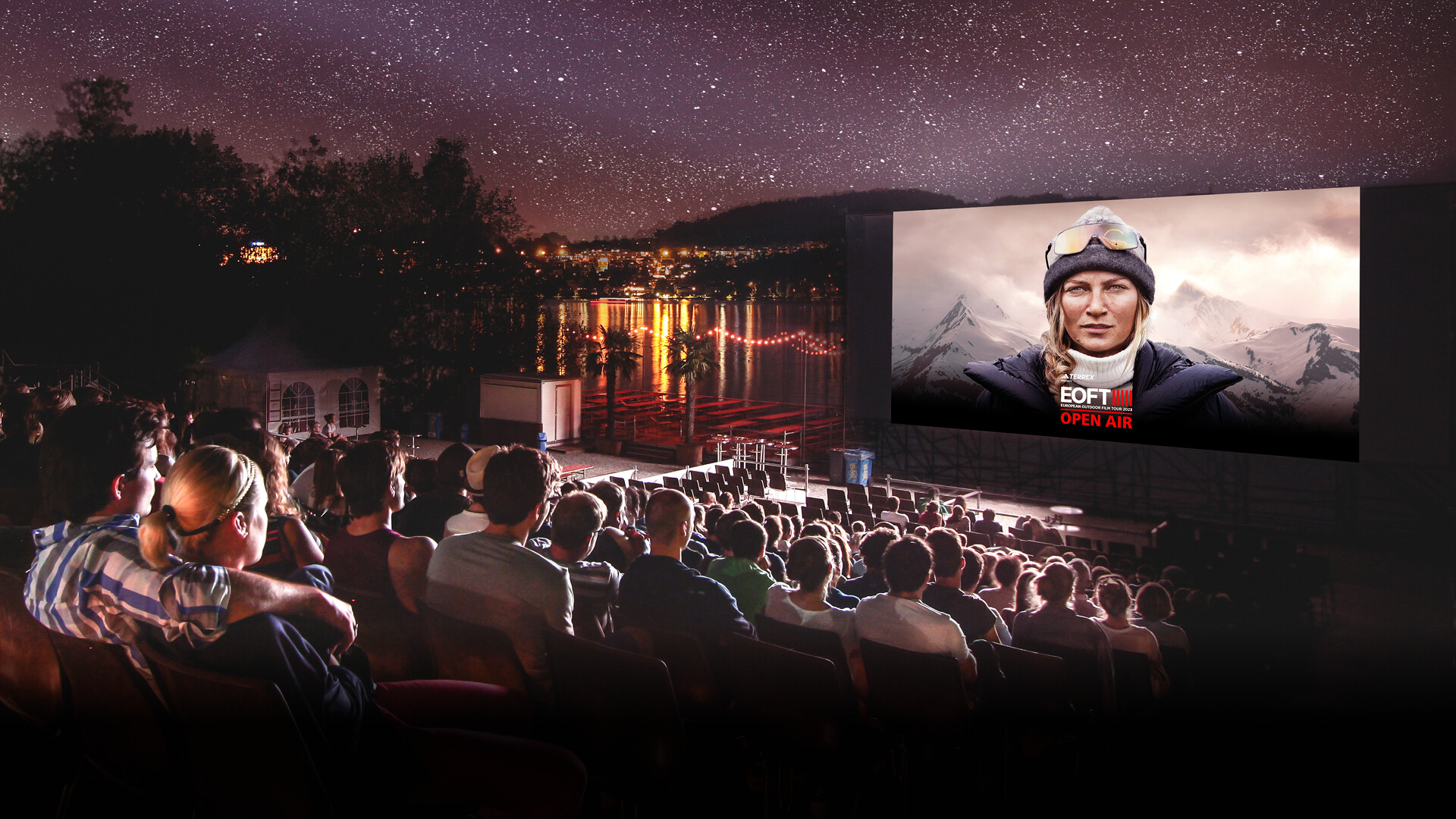 An outdoor cinema by the lake with a large audience watching a film under the starry sky. On the screen is a woman in outdoor gear with mountains in the background, and the text “EOFT OPEN AIR” is displayed.